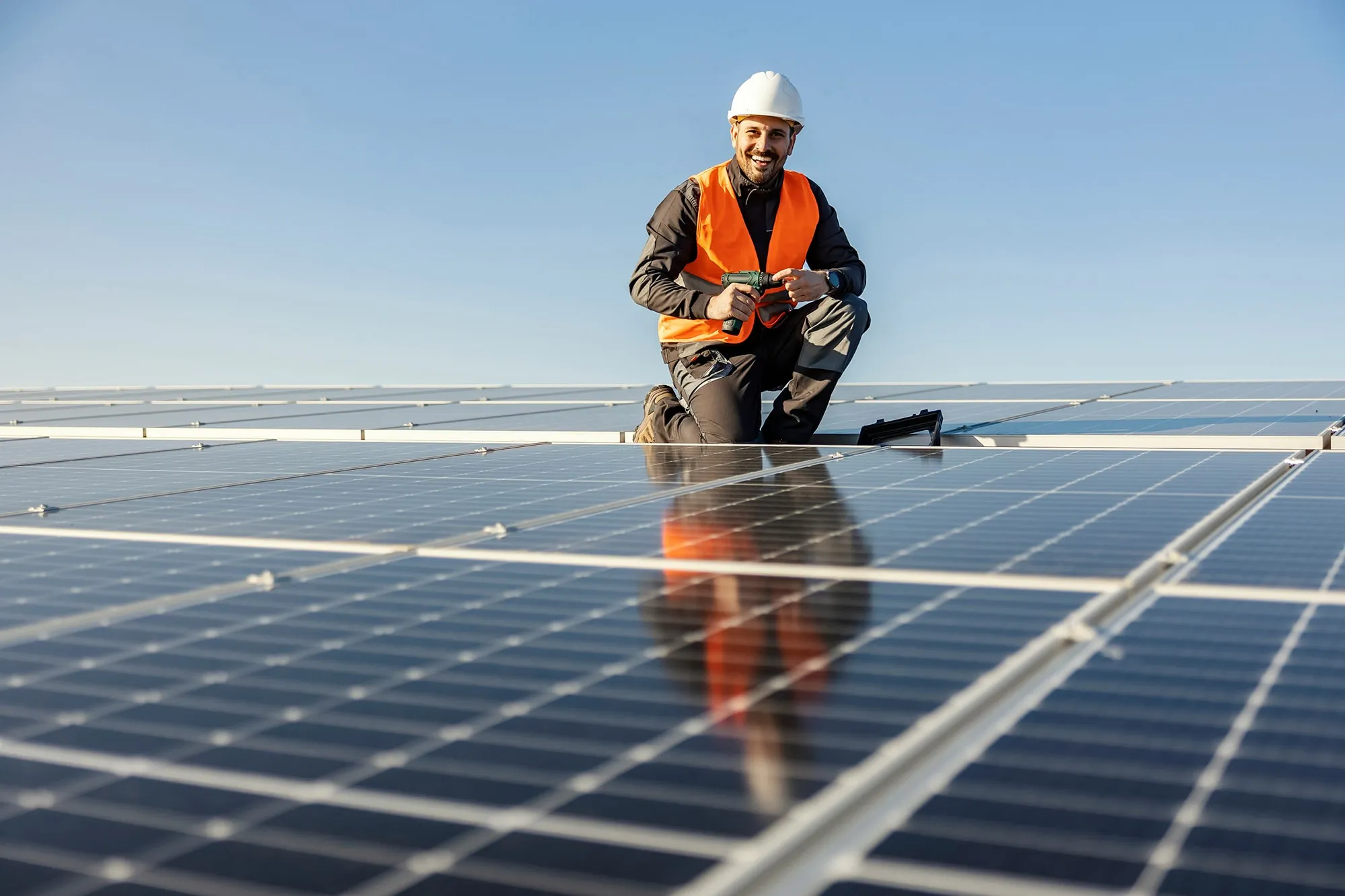Smiling technician wearing a hard hat and orange safety vest kneeling on a solar panel while holding a drill under clear blue sky.